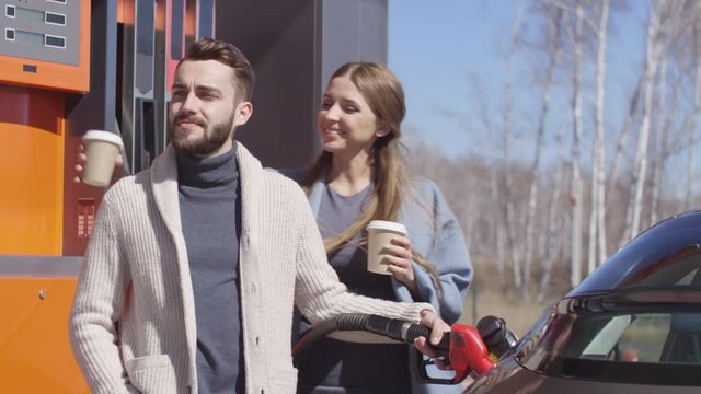 Slow motion shot of man standing at gas station and filling up car while his beautiful girlfriend walking with coffee cups, embracing him and smiling