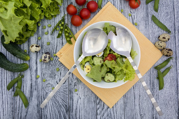 Fresh salad with leaves, tomatoes, cucumber and quail eggs, salad spoon and fork, napkin on grey wooden table