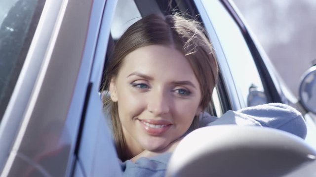 Beautiful young woman sitting on driver seat in car and looking at camera with smile while gas station attendant filling up her car with fuel