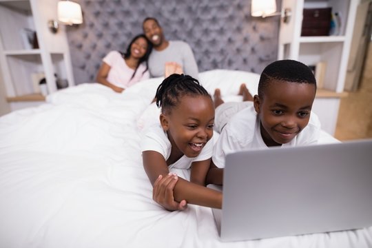 Happy siblings using laptop on bed with parents in background