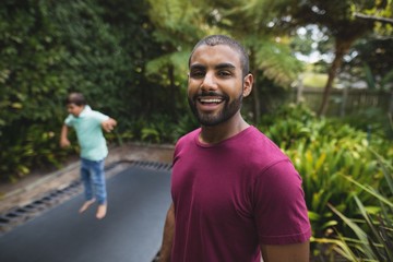 Portrait of smiling father while son jumping on trampoline