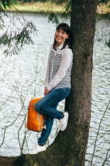 Tourist young smiling girl with orange backpack in hands standing near to the big tree on the bank of the mountain lake surrounded by forest