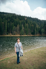 Fototapeta na wymiar Senior beautiful woman standing on the bank of the mountain lake surrounded by forest. Travel concept