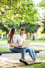 Two female friends in park sitting on bench and working on laptop