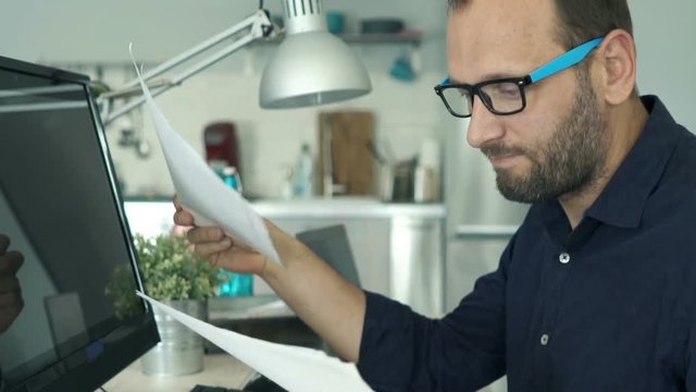 Unhappy, overwhelmed man working with documents and pc computer by table at home
