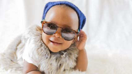 Super cute cool baby. Hipster baby in fur vest and sunglasses lies on a white bed in a room with...
