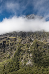 Fiordland National Park, New Zealand - March 16, 2017: A white cloud band hides part of dark rocky mountain peak near Monkey Creek under blue sky. Green vegetation with water flows on mountain slope.