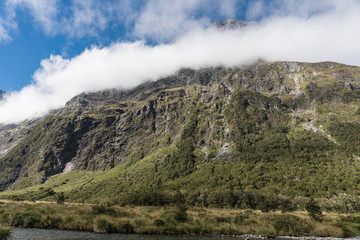 Fiordland National Park, New Zealand - March 16, 2017: Landscape of Monkey Creek flowing in valley among tall mountain peaks partly covered by white cloud bands. Green vegetation in swamp.