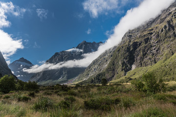 Fiordland National Park, New Zealand - March 16, 2017: Landscape of Monkey Creek flowing in valley among tall mountain peaks partly covered by white cloud bands. Green vegetation in swamp.