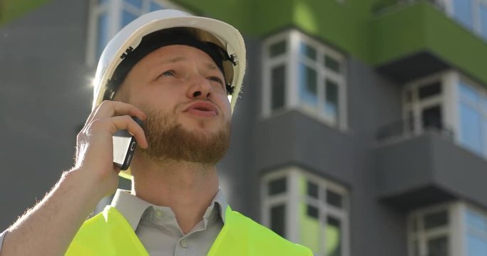 Handsome caucasian builder in white helmet and yellow vest talking on mobile phone on unfinished construction background. Close up.