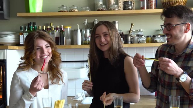 two girls and guy fool around at kitchen with chili pepper and spaghetti