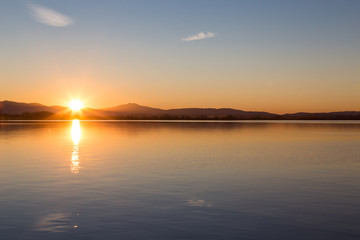 Beautiful sunset at the lake, with two clouds and sun reflecting on water.