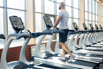 Back view portrait of muscular sportsman running on treadmill listening to music  alone in empty...