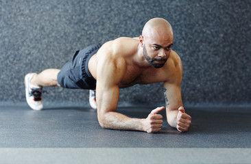 Obraz premium Motivational portrait of shirtless muscular man holding plank against grey background, straining with effort and looking determined during intense endurance workout in gym