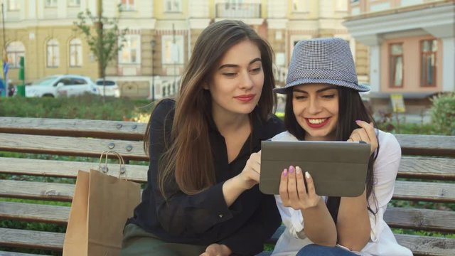 Two pretty female friends watching something on the tablet outdoors. Attractive brunette teenager in gray hat holding gadget in her hand. Cute young brown haired woman pointing her forefinger on the