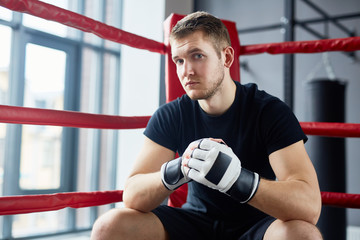 Fototapeta na wymiar Portrait of young man posing in boxing ring sitting on floor and looking at camera looking at camera with confidence