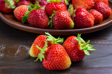 Fresh organic strawberries on dark wooden plank, and full brown plate with strawberry on background, close-up view