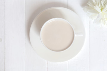 On a white wooden background there is a cup of coffee. Next to it is a white peony flower