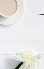 On a white wooden background there is a cup of coffee. Next to it is a white peony flower