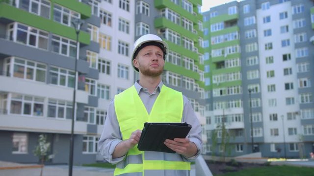 Builder engineer with white protected helmet and safety vest on construction site using tablet for tapping something. Outdoor.