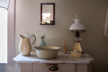 An old wash basin with water jug, wash bowl, soap holder in enamelled metal. Standing on a commode/chest of drawers with a Kerosene lamp and a mirror above