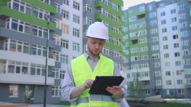 Caucasian builder with white protected helmet and safety vest on construction site using tablet for tapping something. Outdoor.