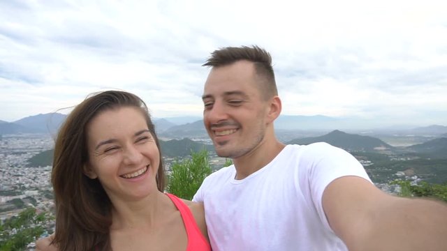 Happy smiling couple recording video, taking selfie in the mountains with aerial city view. Man kisses girl