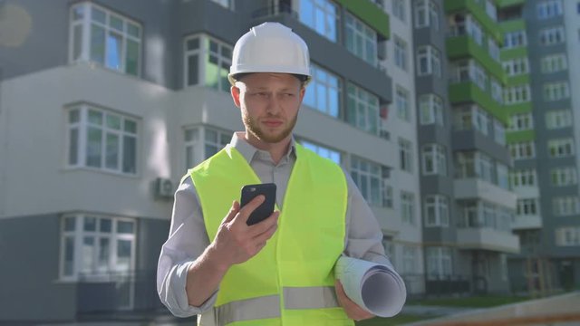 Handsome caucasian worker in white helmet and green vest talking on mobile phone while standing near built house.