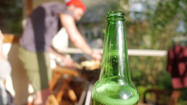 Close up of Green Beer Bottle at Picnic. Young Man Starting Fire on Background.