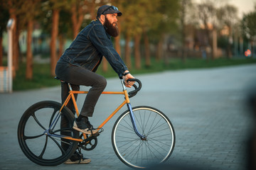 Fototapeta premium Bearded guy and fix bike. Young man with gourgeous large beard in sunglasses and in the cap riding a vintage bicycle in the city park at sunset.