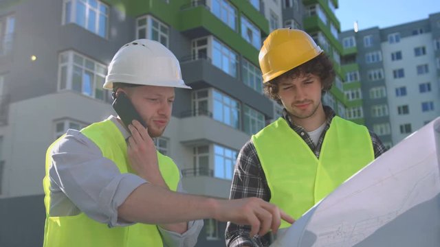 Team of young caucasian builders in protected helmets and vests looking drawing. Builder standing near his collegue using smart phone. Outdoor.
