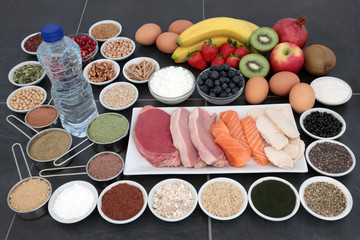Body building health food of high protein lean meat and salmon, supplement powders, fruit, nuts, seeds, grains,  pulses, herbs, dairy and bottled water over slate background.