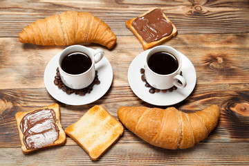 Two cups of coffee, croissant, toast and chocolate on wooden table