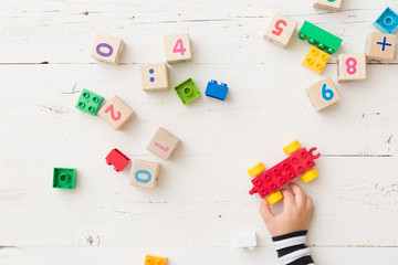 Top view on child's hand playing with wooden cubes with numbers and colorful toy bricks on white wooden background. Baby with toys