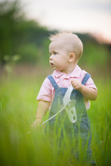 Portrait of cute little blond toddler boy holding paper plane toy on a stick and looking into distance. Adorable child walking in park on sunny day. Summertime Outdoors. Childhood, lifestyle concept