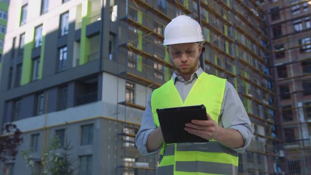 Handsome caucasian builder with white protected helmet and green safety vest standing at unfinished construction and reporting his work using tablet. Outdoor.
