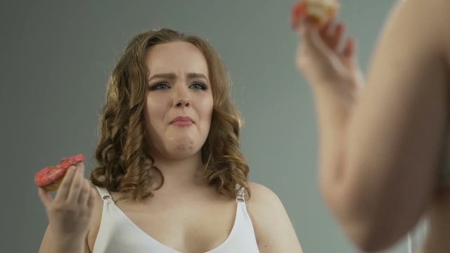 Depressed overweight girl looking at body in mirror, eating donut in despair