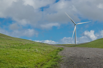 A gravel road leads to a wind turbine on a hillside, against the backdrop of cloud-filled blue sky in the Livermore, California wind farm