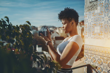 Charming young afro american lady shooting on her vintage film photo camera views of Lisbon from balcony, attractive pensive Brazilian teenage girl making photos on her retro camera from high point