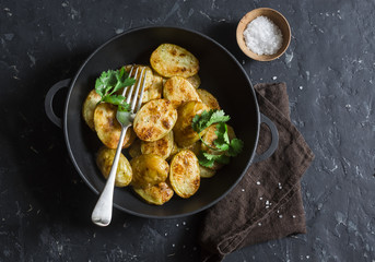 Baked new potatoes with coriander in a cast iron skillet on the dark table, top view. Vegetarian lunch or snack
