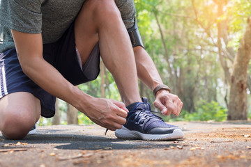 Cropped shot of young man runner tightening running shoe laces, getting ready for jogging exercise outdoors. Male jogger lacing his sneakers standing on forest path before morning run