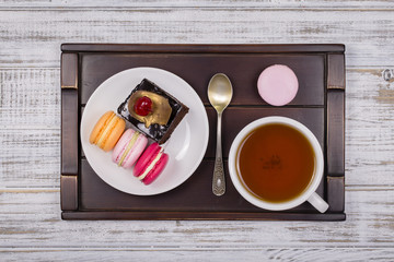 Cup of tea, piece chocolate cake and macaron cakes on tray on white wooden table. Lifestyle concept.