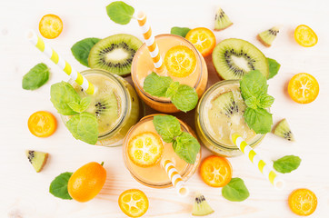 Freshly blended orange kumquat and green kiwi fruit smoothie in glass jars with straw, mint leaf, cut ripe berry, top view, close up. White wooden board background.