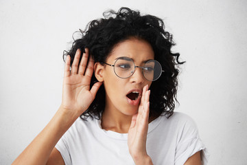 Curious African woman with curly hair wearing glasses and white top holding hand near her ear...