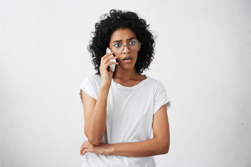 Fototapeta na wymiar Afro American housewife with curly hair wearing big round glasses and casual T-shirt talking over smartphone with her friend while having free time having surprised look after hearing shocked news