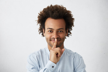 Happy dark-skinned guy with African hairstyle wearing elegant white shirt showing silence sign asking to be quiet. Indoor portrait of mixed race man holding his finger on lips having pleasant look - obrazy, fototapety, plakaty