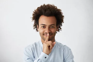 Foto op Plexiglas Happy dark-skinned guy with African hairstyle wearing elegant white shirt showing silence sign asking to be quiet. Indoor portrait of mixed race man holding his finger on lips having pleasant look © Wayhome Studio
