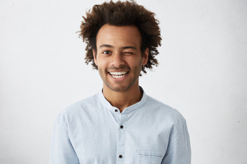 Funny African man with bushy curly hair blinking his eye having warm broad smile wearing white...