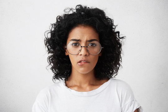 Headshot of African pretty female with dark eyes, well-marked eyebrows and bushy curly hairstyle wearing big round glasses and white T-shirt biting her lip while having self-confidence and doubt
