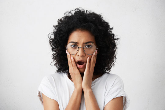 Waist up portrait of fearful mixed race woman with short curly dark hair wearing big round shades and T-shirt holding hands on cheeks looking with opened mouth into camera having shok and disbelief
